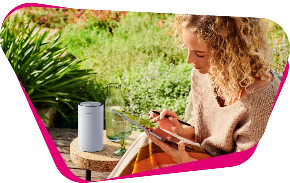 Eine Frau sitzt im Garten, nutzt ein Tablet mit einem Stift. Auf dem Tisch stehen eine Glasflasche, ein Glas und eine weiße, zylinderförmige Speedbox.